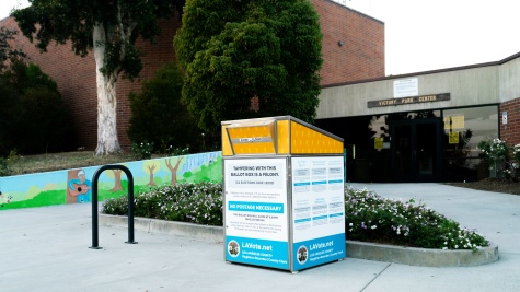 An image of a red, white, and blue ballot drop box in front of a civic building.