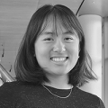 A young woman with a shoulder-length bob and bangs smiles at the camera. She is wearing a black shirt and a simple chain necklace.