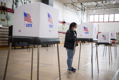 A woman of color, wearing a mask, jacket, and jeans, stands at a voting booth and fills out her ballot.