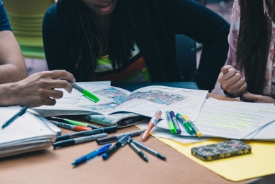 A table is covered in colored pens and markers. Three students are looking at notebooks and papers spread out in front of them. One student on the left is pointing to a point on a map in front of them with a highlighter.